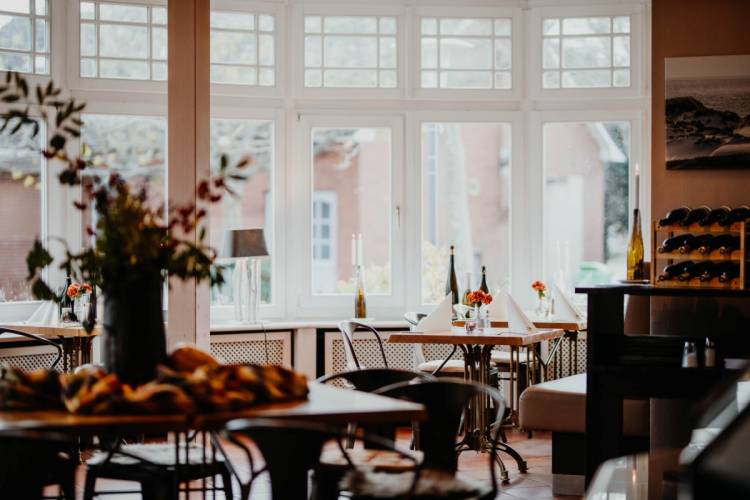 Elegantes Restaurant im Hotel Hüttmanns auf Amrum mit gedeckten Tischen, großen Fenstern, Weinfalschen und Dekoration.