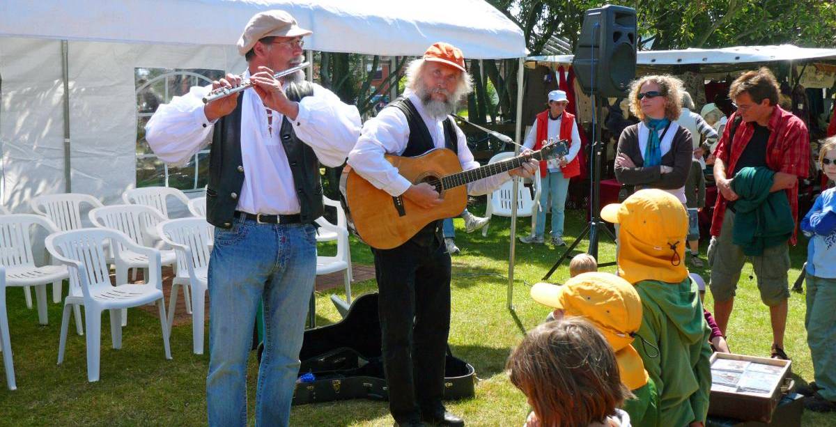 Zwei Musiker mit Flöte und Gitarre spielen auf einem Sommerfest vor Kindern und Erwachsenen unter freiem Himmel