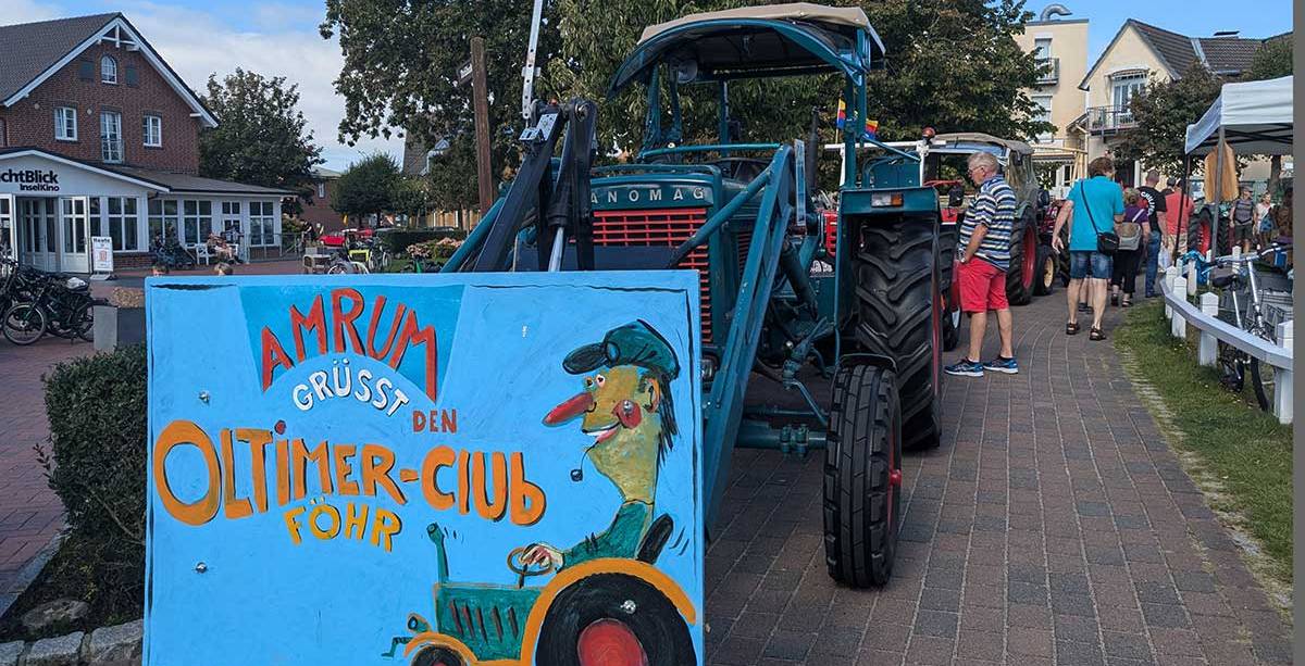 Ein Hanomag-Traktor mit bemaltem Schild „Amrum grüßt den Oldtimer-Club Föhr“ mit Besuchern.